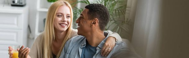 smiling blonde woman holding orange juice and hugging african american boyfriend in kitchen, banner - Photo, image