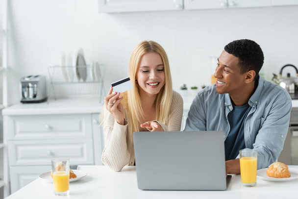 happy young woman holding credit card and pointing at laptop near smiling african american boyfriend and breakfast in kitchen - Foto, imagen