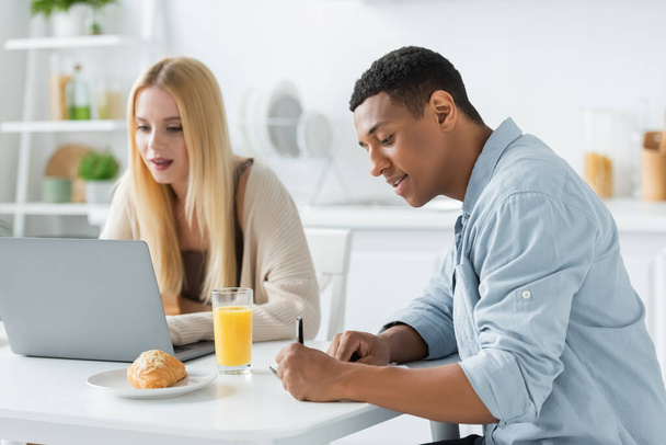 hombre afroamericano escribiendo en cuaderno y mujer rubia trabajando en el ordenador portátil cerca del desayuno en la cocina - Foto, imagen