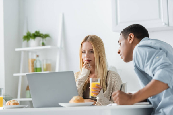 femme réfléchie avec du jus d'orange regardant ordinateur portable près flou copain afro-américain dans la cuisine - Photo, image
