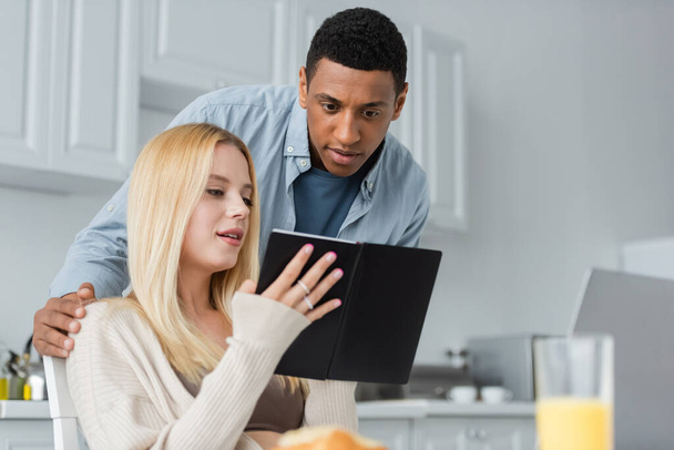blonde woman showing copybook to african american boyfriend while working in kitchen - Foto, immagini