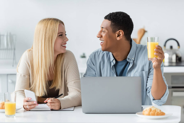 happy interracial couple looking at each during breakfast near laptop and notebook  - Foto, Bild
