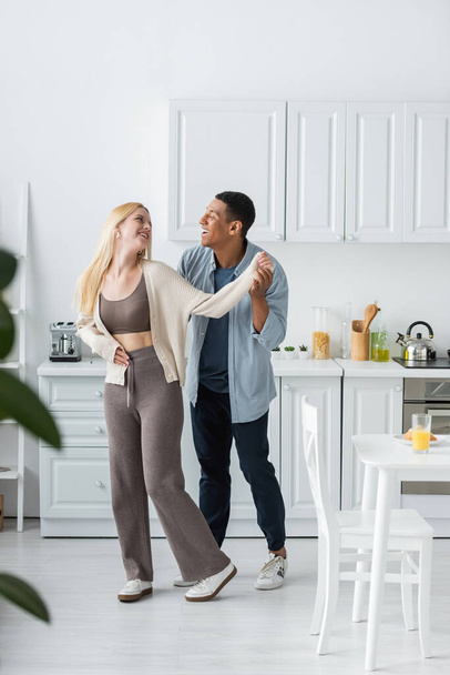 full length of joyful interracial couple smiling at each other while dancing in modern kitchen - Foto, Imagem