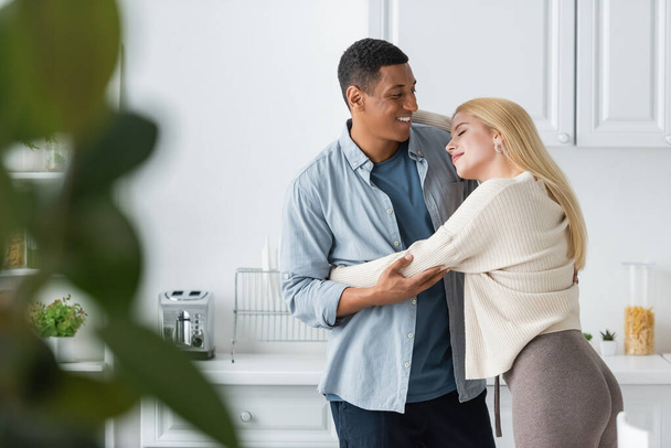 happy african american man hugging young blonde girlfriend on blurred foreground in kitchen - Фото, изображение