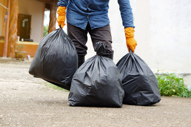 Closeup man holds black plastic bag that contains garbage inside, stand in front of house. Concept : Waste management. Environment problems. Daily chores. Throw away rubbish .              - 写真・画像