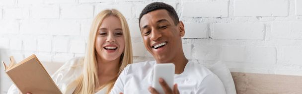vrolijk Afrikaans amerikaans man tonen smartphone naar lachende vriendin holding boek in slaapkamer, banner - Foto, afbeelding