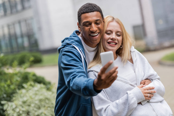 cheerful interracial couple in hoodies taking selfie on mobile phone outdoors - Fotó, kép