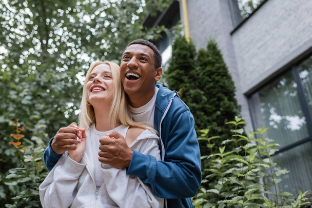 excited african american man holding hands of blonde girlfriend and laughing on street - Fotografie, Obrázek