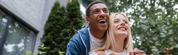 low angle view of excited african american man laughing near blonde girlfriend on city street, banner - Zdjęcie, obraz
