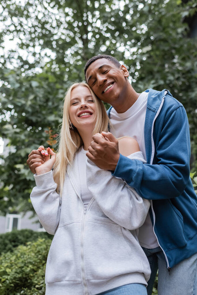 low angle view of pleased african american man with closed eyes holding hands of young girlfriend outdoors - Фото, изображение