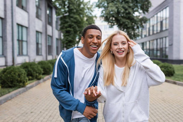 happy multiethnic couple in hoodies holding hands and looking at camera on urban street - Valokuva, kuva