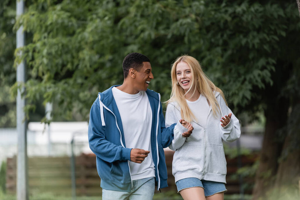 excited blonde woman gesturing while talking to african american boyfriend in park - Photo, image