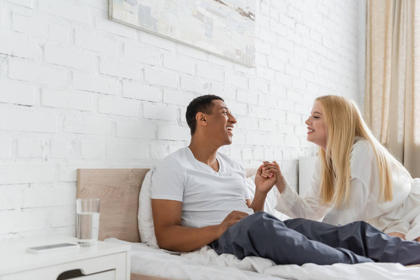 joyful multiethnic couple holding hands and looking at each other on bed - Photo, Image