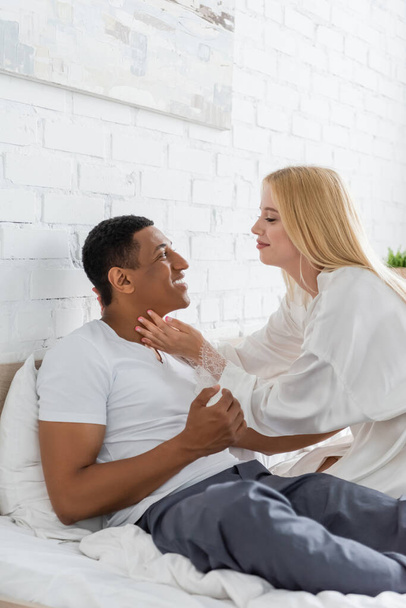 side view of blonde woman in white robe touching neck of african american boyfriend smiling on bed - Photo, Image