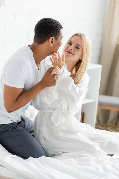 mujer sonriente en túnica blanca tocando la cara del novio afroamericano sosteniendo su mano en el dormitorio - Foto, Imagen