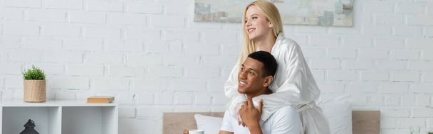 young blonde woman in white robe embracing african american man and looking away in bedroom, banner - Photo, image