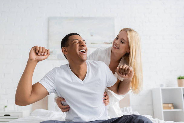 cheerful woman tickling african american boyfriend laughing in bedroom - Photo, Image