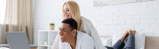 young blonde woman and cheerful african american man watching comedy on laptop in bedroom, banner - Фото, изображение
