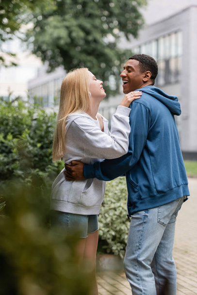 side view of laughing african american man and blonde woman embracing and looking at each other on street - Zdjęcie, obraz