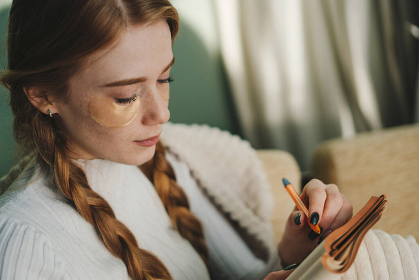 Mujer joven y atractiva con ojos haciendo rutina matutina de cuidado facial mientras toma notas en su diario. Mantener un diario - Foto, Imagen