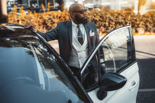 View of a handsome bald black man entrepreneur with a fine black beard, in sunglasses and a tailored fashionable suit, looking aside while entering his contemporary white car parked outdoors - Photo, Image