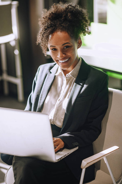 Een vrolijke bui. vrij jong volwassen mulatta vrouw werken op laptop glimlachen op camera zitten op stoel in kantoor - Foto, afbeelding