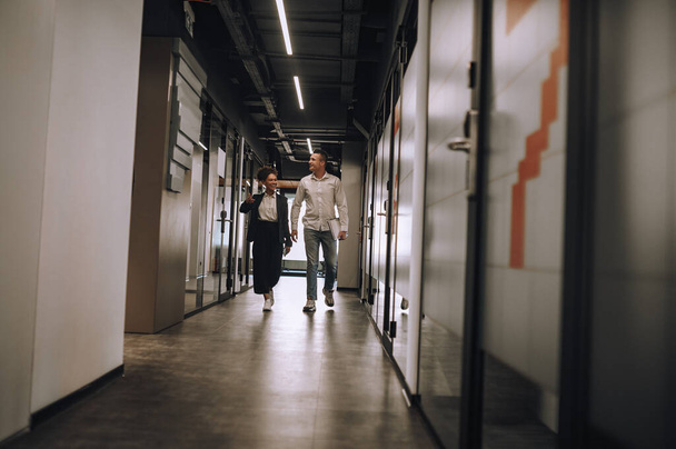 Colleagues. Caucasian young adult man and mulatta woman talking walking in illuminated office corridor - Foto, imagen