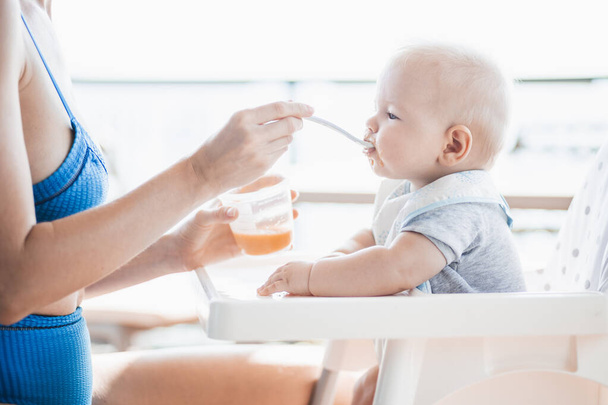 Mother spoon feeding her baby boy child in baby chair with fruit puree on a porch on summer vacations. Baby solid food introduction concept - Photo, Image