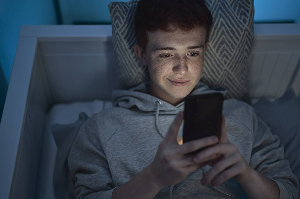 Cheerful caucasian teenage boy using mobile phone while lying in bed at night in his room  - Photo, Image
