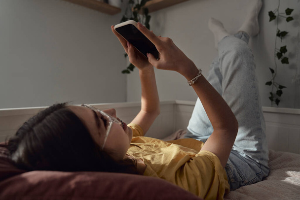 Caucasien adolescent fille navigation téléphone tout allongé sur le lit avec ses jambes vers le haut - Photo, image