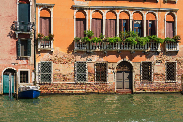 ヴェネツィア運河沿いのヴィンテージの建物のファサード。窓のシャッター、古い家のバルコニーの花、ヴェネツィア通りの家の入り口のボート、イタリアの美学 - 写真・画像