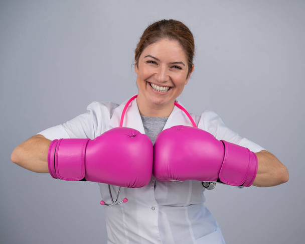 Smiling female doctor in pink boxing gloves with a pink ribbon on a gray background. Fight against breast cancer - Zdjęcie, obraz