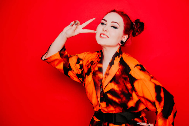 Beautiful girl on a red background. Red hair and tunnels in the ears, red dress and lips. Anime poses. The girl with eyeliner and two buns on her head grimaces, smiles, has fun. Peace gesture - Fotó, kép