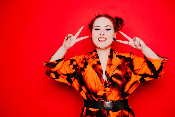 Beautiful girl on a red background. Red hair and tunnels in the ears, red dress and lips. Anime poses. The girl with eyeliner and two buns on her head grimaces, smiles, has fun. Peace gesture - Фото, изображение