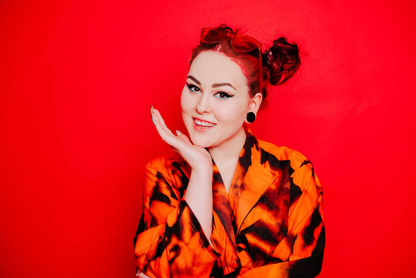 Cute girl on a red background. Red hair and tunnels in the ears, red dress and lips. Anime poses. The girl with eyeliner and two buns on her head grimaces, smiles, has fun. Japanese culture - Fotó, kép