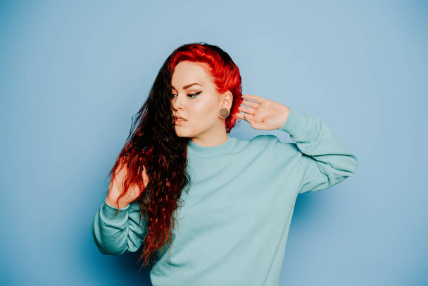 Woman in a blue sweater on a blue background posing. A beautiful brunette with long hair and and tunnels in her ears. Eyeliner and trendy makeup - Photo, Image