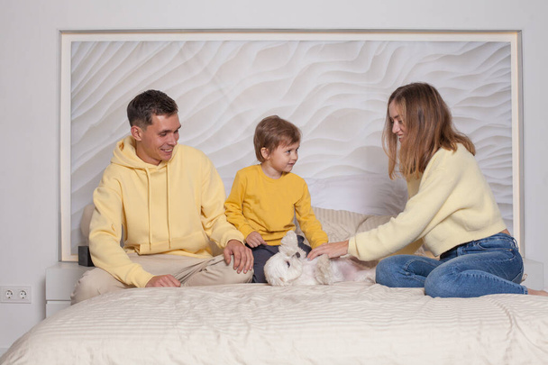 Υπέροχη οικογένεια, χαριτωμένοι γονείς, μαμά και μπαμπάς με αξιολάτρευτο παιδί γιος κάθεται στο κρεβάτι και παίζει με το μικρό λευκό σκυλί - Φωτογραφία, εικόνα