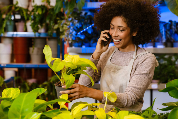 Молодая счастливая девушка-флористка разговаривает по телефону во время работы с цветочными горшками в цветочном магазине - Фото, изображение