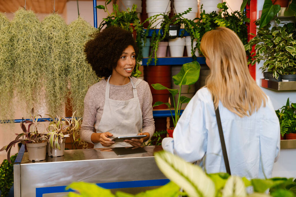 African american florist girl standing at cash register while working with female customer in flower shop - Foto, Imagen