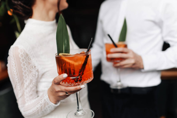 De handen van vrouw en man kloppen, proost met een glas Spritz cocktail. Echtpaar vieren bruiloft, jubileum met Aperol spritz cocktails, met sinaasappel en groen Verfrissende alcoholische drank. - Foto, afbeelding
