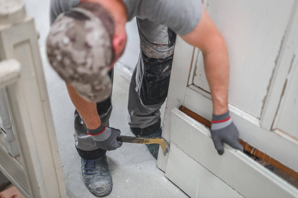 A young caucasian man in a uniform and gray textile gloves using a crowbar dismantles an old wooden door while standing in an inclination in a room where repairs are underway, side view close-up with selective focus.Construction work concept. - 写真・画像