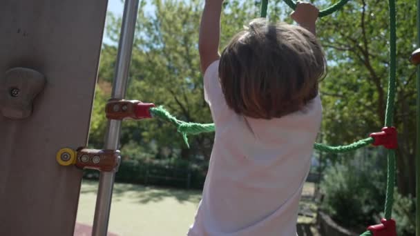 Een actief kind houdt zich vast in een touwladder op de speelplaats buiten in het stadspark. Klein kind klimmen speeltuin structuur buiten - Video
