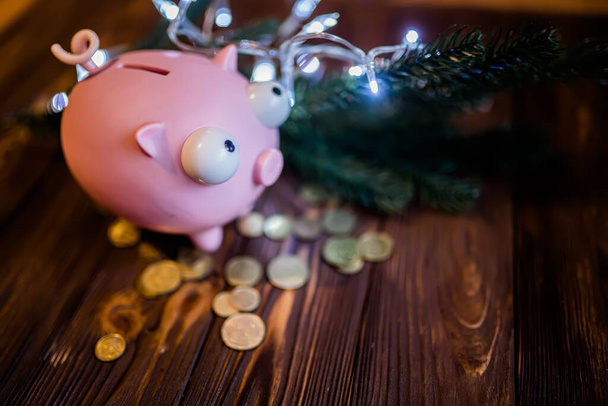 Piggy τράπεζα σε ένα διπλό τραπέζι αγρόκτημα με διάσπαρτα νομίσματα και γιρλάντες με φώτα. Χριστουγεννιάτικη διακόσμηση. Περνάω τις πρωτοχρονιάτικες διακοπές. Αποεστίαση - Φωτογραφία, εικόνα