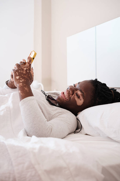 Young African American woman with vitiligo lying in comfortable bed under blanket using cellphone surfing internet enjoying free time - Photo, Image