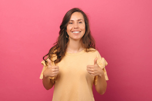 Una joven sonriente sostiene dos pulgares hacia arriba por una recomendación. Stdusio rodó sobre fondo rosa. - Foto, imagen