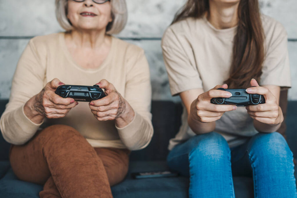 Foto de mamá e hija mayores graciosas sentadas cómodas en el sofá, sosteniendo el joystick jugando videojuegos, pasando el fin de semana juntas. Estilo de vida. Ordenador - Foto, Imagen