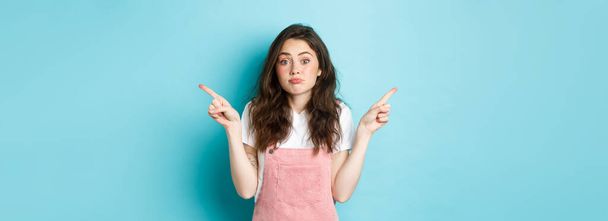 Clueless and confused cute timid girl, shrugging unaware, pointing fingers sideways at two sides with indecisive face, standing over blue background. - Photo, Image