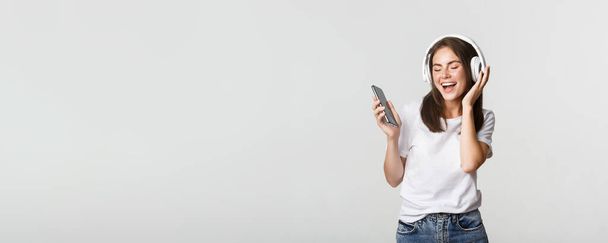Happy beautiful brunette girl dancing and listening music in wireless headphones, holding smartphone. - Photo, Image