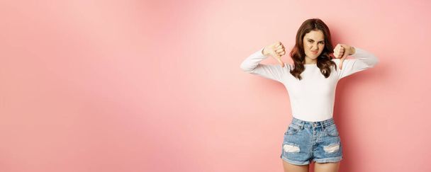 Portrait of young woman showing thumbs down to express dislike, disappointed, standing over pink background. - Photo, Image