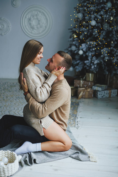 A couple in love on New Year's Eve in a cozy home environment near the Christmas tree. New Year's love story. Beautiful couple in sweaters posing in studio. A guy and a girl celebrate the new year - Фото, изображение